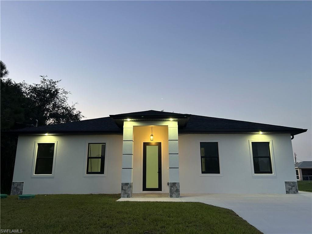 Stucco exterior with a black shingle roof, featuring a covered entry, decorative stone accents, and exterior lighting