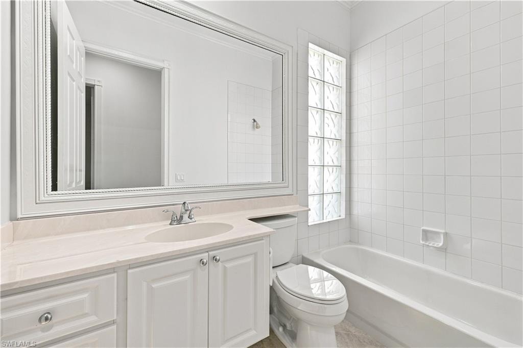 White-tiled bathroom featuring a glass block window, built-in bathtub, and vanity with an integrated sink and light-toned countertop