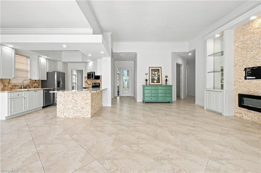 Open-concept kitchen with white cabinetry and stainless steel appliances, central island with textured stone finish, tile flooring, stone-clad fireplace with integrated media niche, and built-in display cabinetry