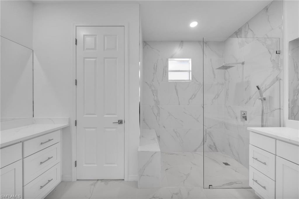 Spacious bathroom featuring a frameless glass shower enclosure with a rainfall showerhead, dual vanities with white cabinetry and light countertops, and large format marble-look tile flooring and shower surround