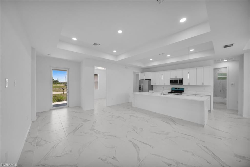 Open-concept living space featuring large format marble-look tile flooring, a spacious kitchen island with integrated sink, white cabinetry, stainless steel appliances, and tray ceilings with recessed lighting