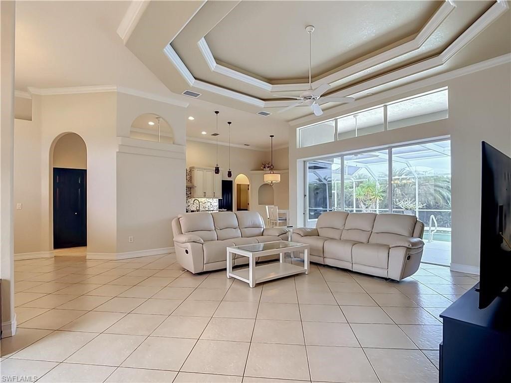 Tray ceiling with crown molding, ceiling fan, tile flooring, arched doorways, and sliding glass doors to the enclosed pool area