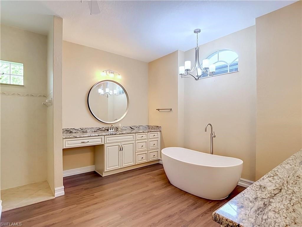 Freestanding soaking tub, wood-finish flooring, dual vanity with granite countertop, round vanity mirror, and a walk-in shower