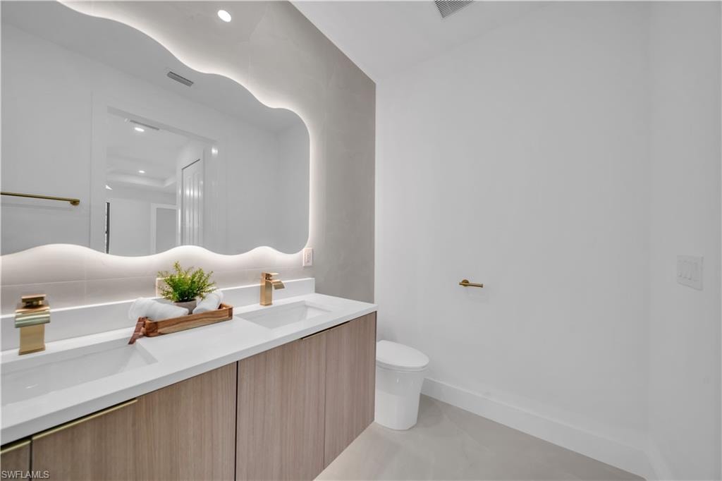 Floating vanity with light wood-finish cabinetry, dual undermount sinks with brushed brass fixtures, and a large illuminated mirror