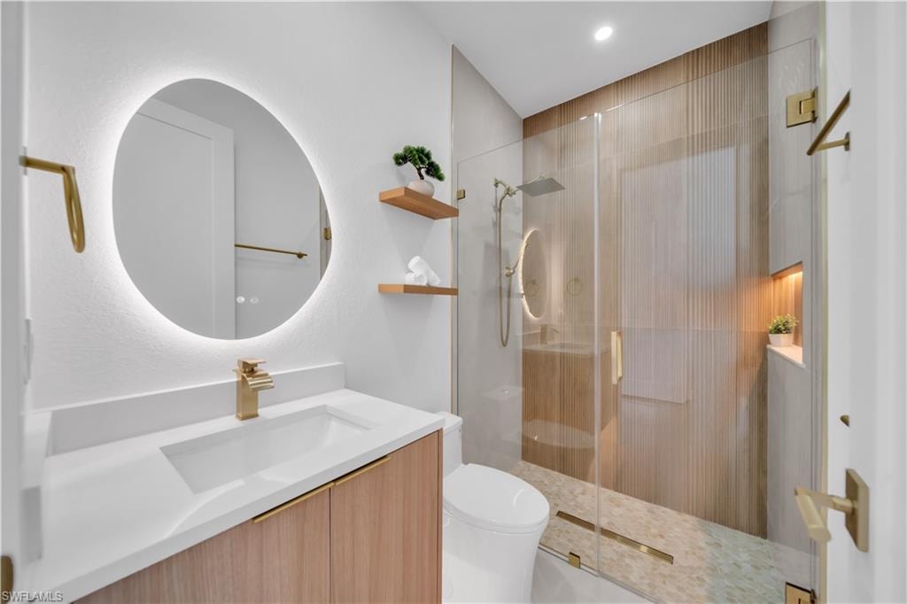 Backlit oval mirror, floating wood-finish vanity with an integrated sink, glass-enclosed shower with a rain shower head, and an illuminated shower niche