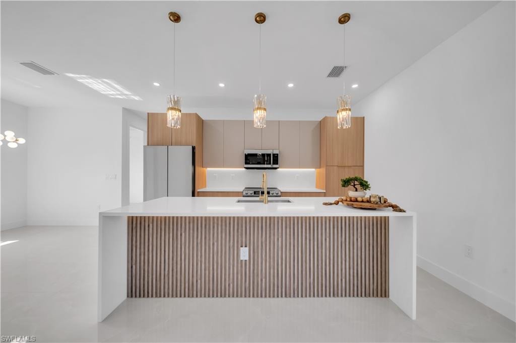Kitchen with white countertop island, wood-slat island accent, gold-toned pendant lighting, light wood cabinetry, and stainless steel appliances