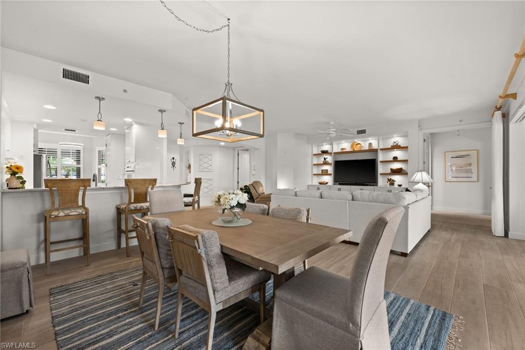 Open-concept living space featuring wood-finish flooring, a built-in media center with shelving, a kitchen island with pendant lighting, and a linear chandelier in the dining area