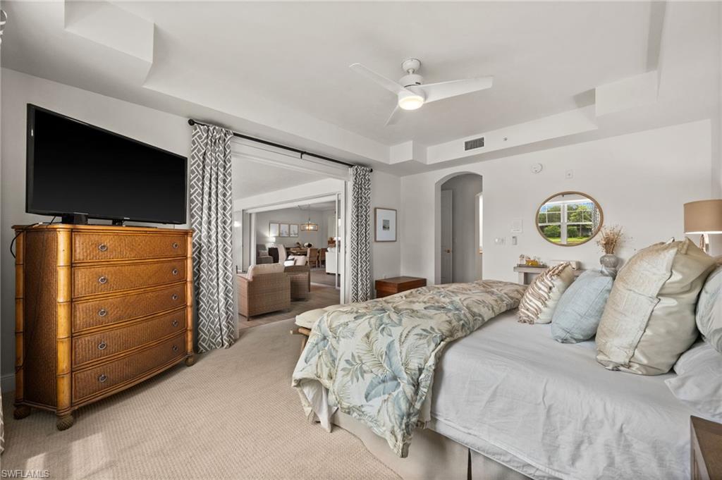 Spacious primary bedroom suite featuring coffered ceiling detail, wall-mounted flat-screen television, wood-finish dresser with multiple drawers, ceiling fan with remote control, carpeted flooring, and arched doorway opening to adjoining sitting area