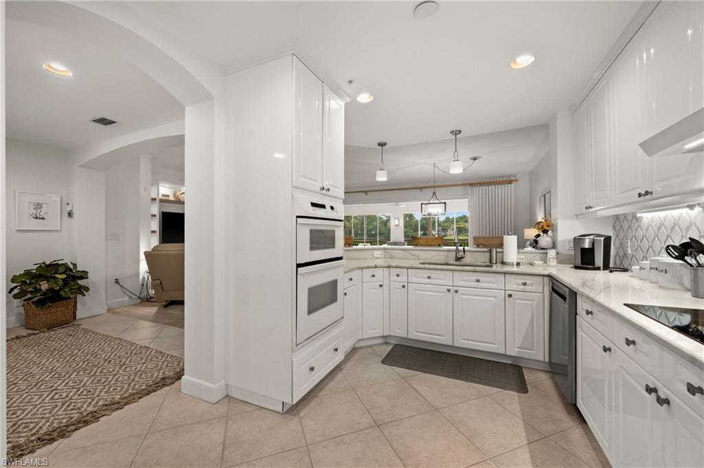 Recessed lighting illuminates a kitchen featuring white cabinetry, stone countertops, integrated ovens, and a tile backsplash