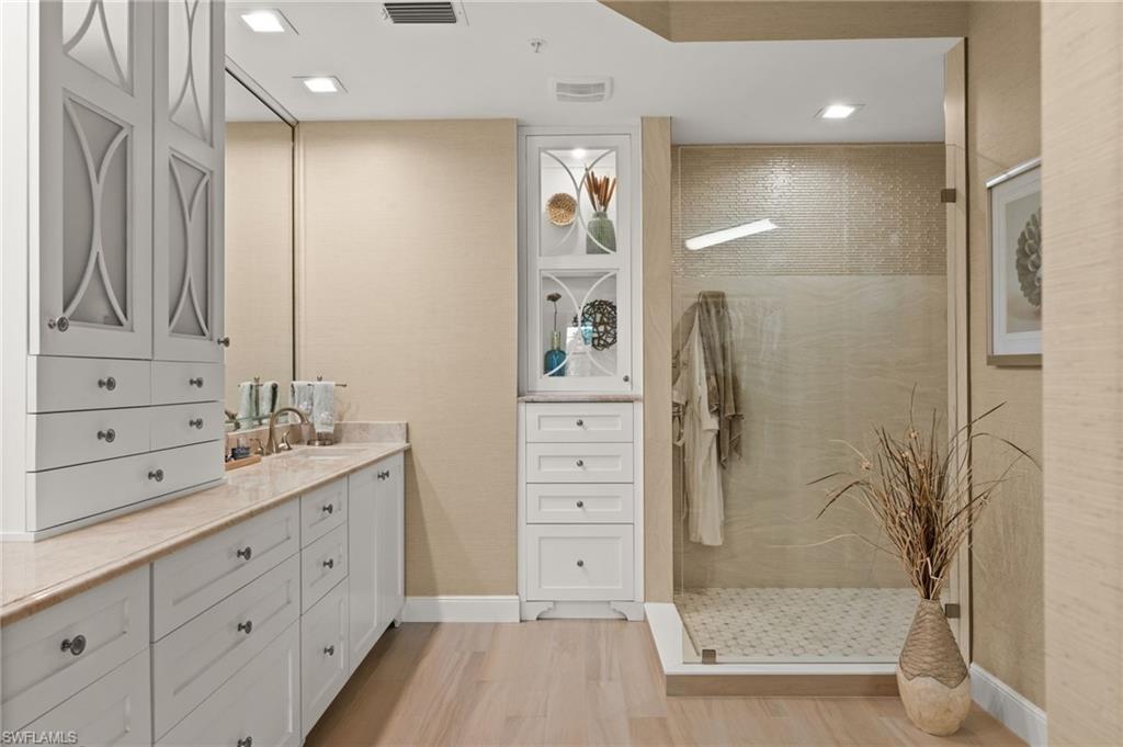 Dual vanity with white cabinetry and stone countertop, glass-enclosed shower with tiled walls, wood-finish flooring, recessed lighting, and built-in storage