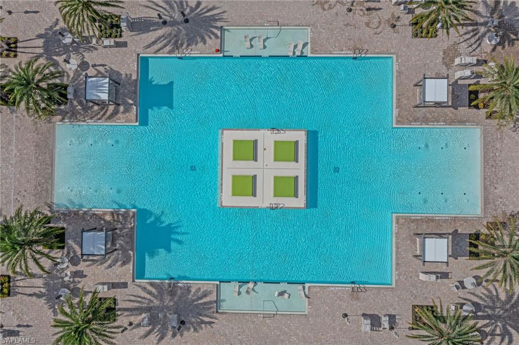 Expansive cross-shaped swimming pool with a central lounging platform, surrounded by a paver deck, multiple cabanas, and mature palm trees
