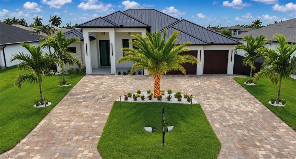 Contemporary architecture featuring a dark metal roof, a white stucco exterior, an extended paver driveway, and a manicured lawn with mature palm trees