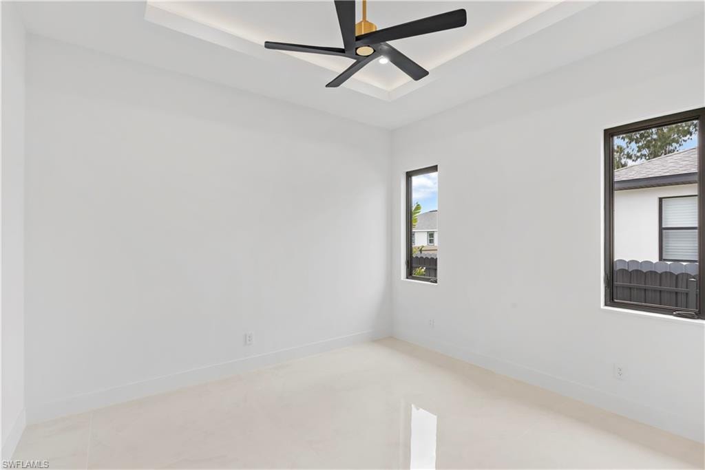 Tray ceiling with recessed lighting, ceiling fan with integrated light, two windows with dark frames, and light-toned tile flooring
