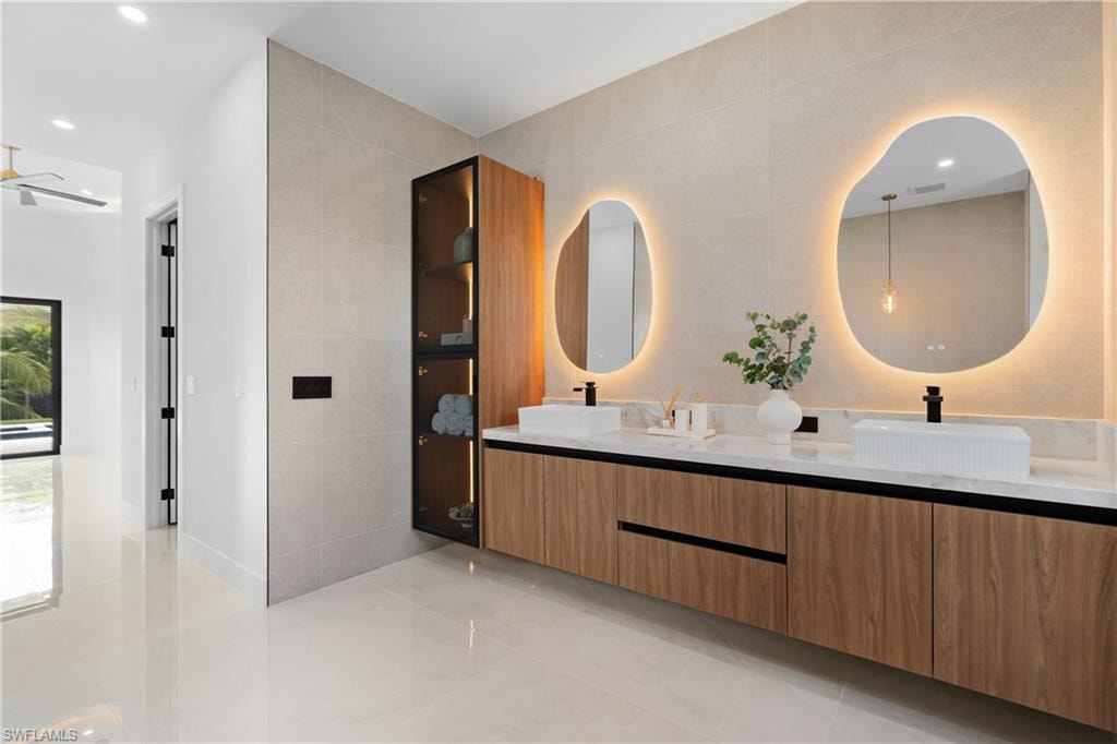 Dual vessel sinks with individual backlit irregular mirrors, wood-finish vanity with integrated storage, and a floor-to-ceiling illuminated cabinet