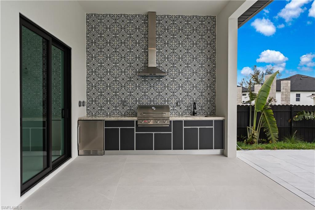 Outdoor kitchen featuring a built-in grill, stainless steel refrigerator, black cabinetry with light countertops, and a patterned tile backsplash