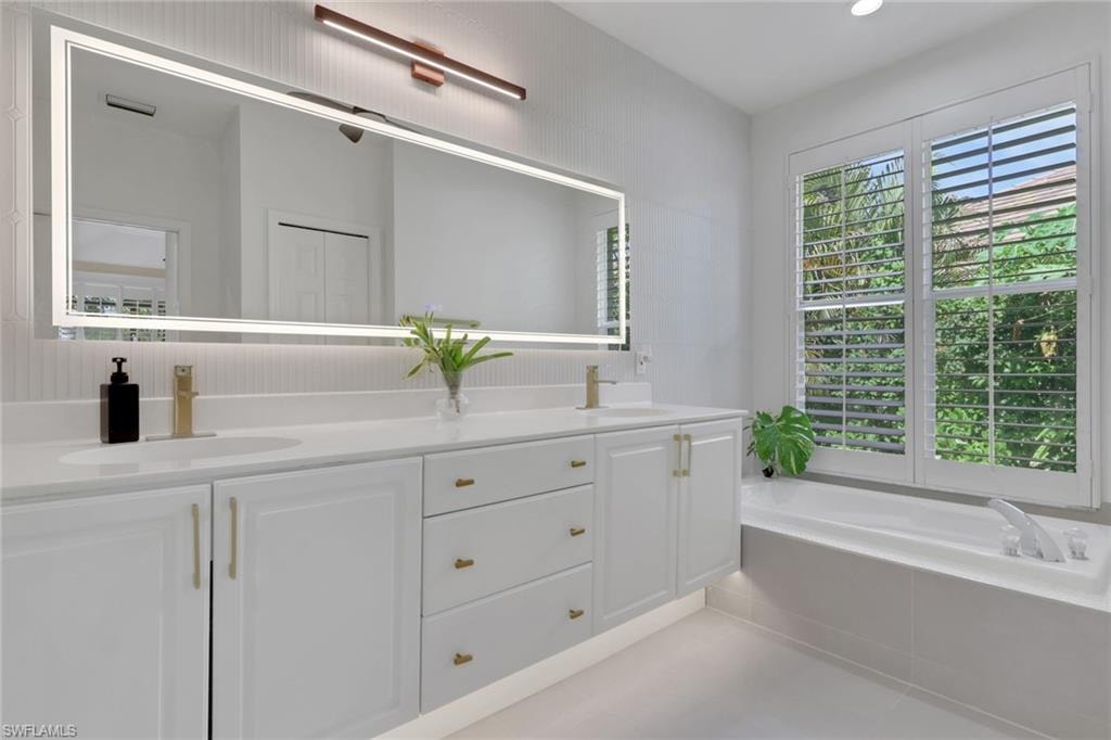 Dual vanity with integrated sinks, white cabinetry, gold-finish hardware, backlit mirror, and a built-in soaking tub with plantation shutters