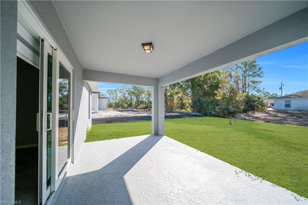 Covered patio with textured concrete flooring, sliding glass doors, and a ceiling-mounted light fixture, opening to a grass lawn