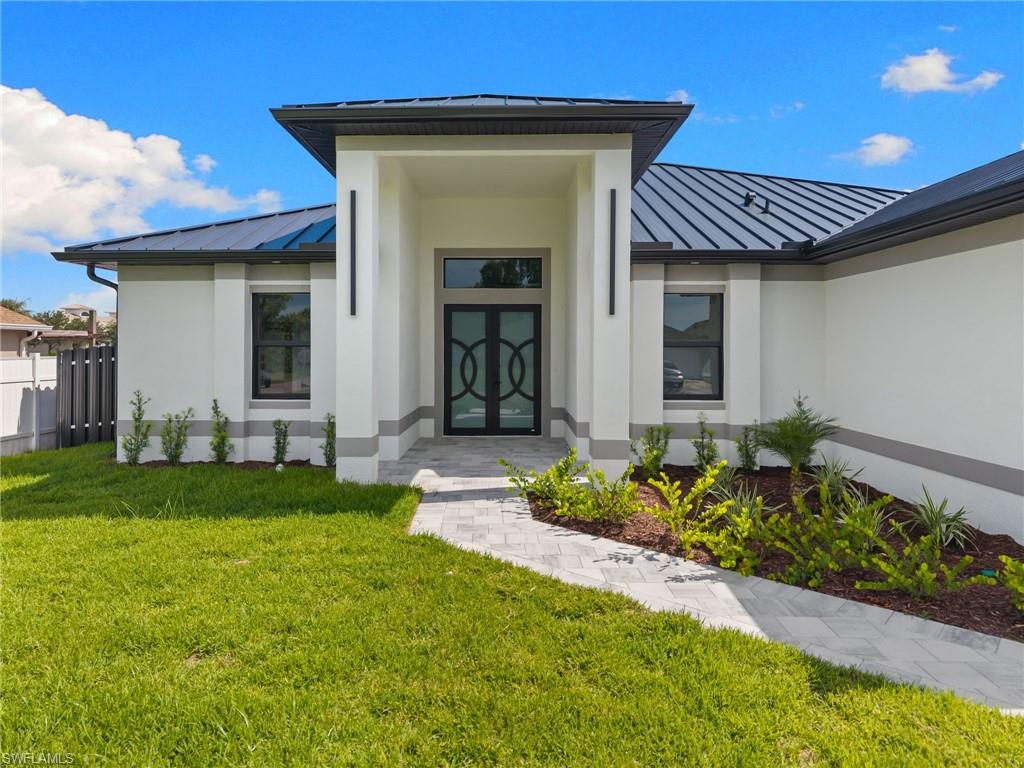 Stucco exterior with a dark metal roof, featuring a paver walkway leading to a double-door entry with decorative glass