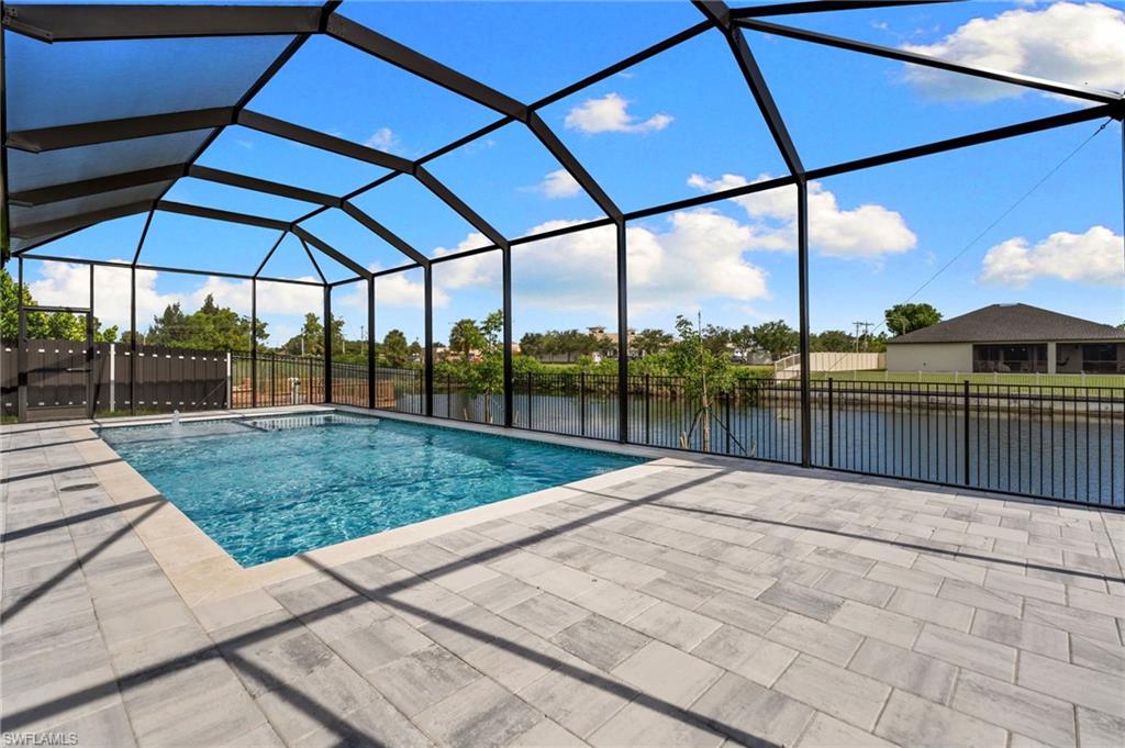 Screened-in pool enclosure featuring a rectangular pool with water features, a spacious paver patio, and a metal fence bordering a canal