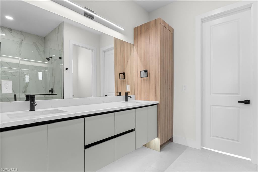 Dual vanity with integrated sinks, matte black fixtures, full-width mirror, light wood-finish cabinetry, and a glass-enclosed shower