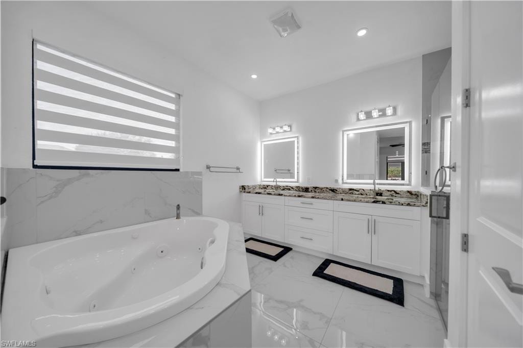 Soaking tub with surrounding white marble-look tile, dual vanity with granite-look countertops, and illuminated mirrors