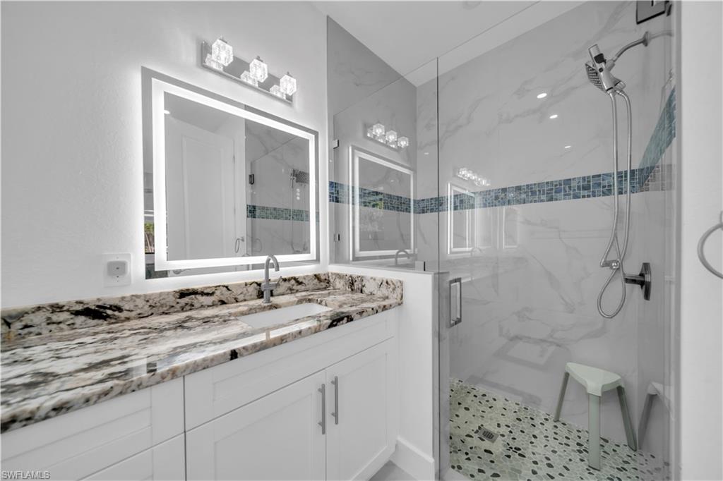 Spa-inspired primary bathroom featuring a large-format marble-look tile shower with rainfall showerhead and hand shower, illuminated vanity mirror with integrated LED lighting, dual-sink vanity with patterned stone countertop, and decorative tile accent band