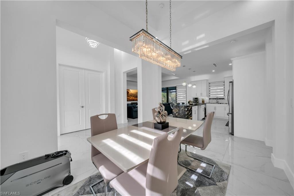 Open-concept dining area with a linear crystal chandelier, light-toned flooring, and direct access to a modern kitchen featuring a dark-toned island with light veining
