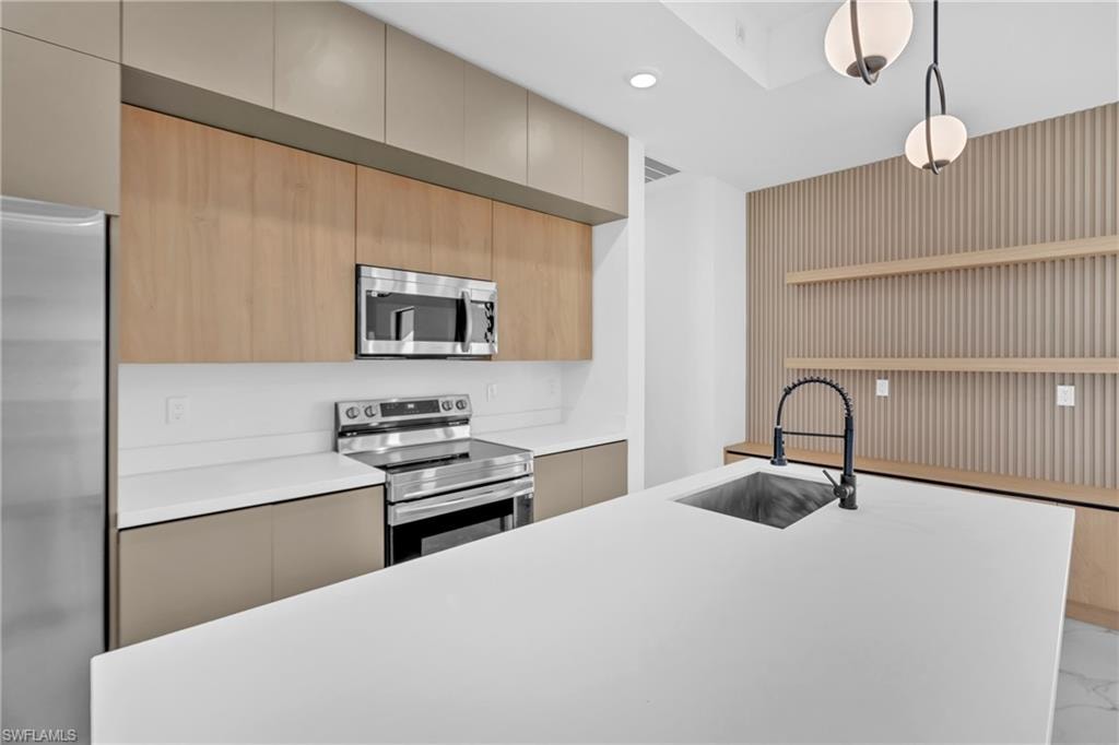 Kitchen featuring stainless steel appliances, wood-finish cabinetry, a white countertop island with an undermount sink, and decorative slatted wall paneling with floating shelves