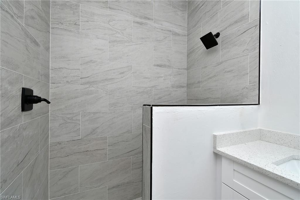 Tiled shower with matte black fixtures, glass shower partition, white vanity with shaker-style door, and speckled countertop