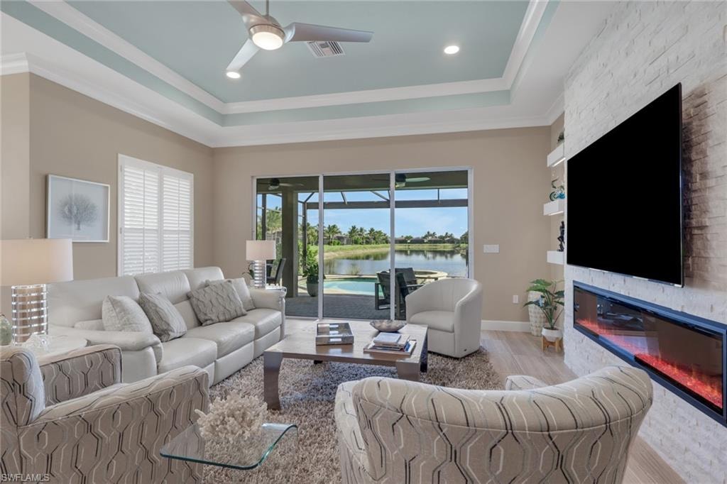 Tray ceiling with recessed lighting and a ceiling fan, sliding glass doors, white brick-patterned accent wall with an inset linear fireplace, plantation shutters, and wood-finish flooring