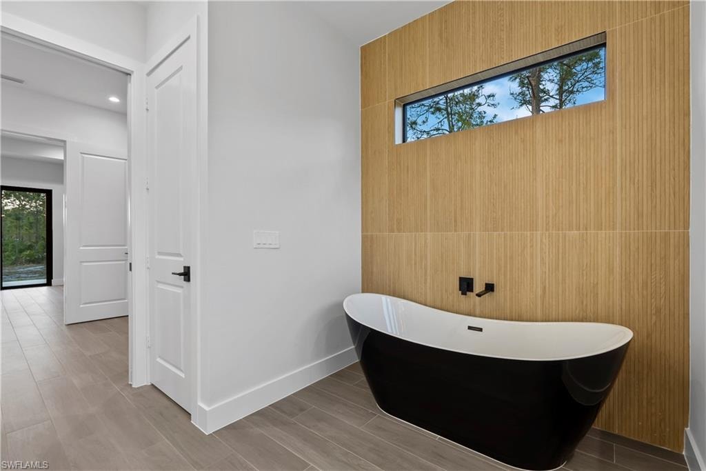Freestanding black and white soaking tub, wood-finish wall paneling, horizontal transom window, matte black wall-mounted bath filler, and wood-finish flooring