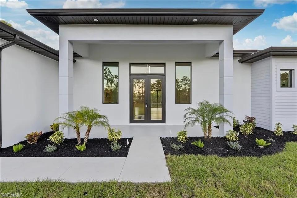 Modern architectural facade featuring a covered entry, dark-framed double entry doors, large sidelight windows, and landscaped planting beds with mature palms