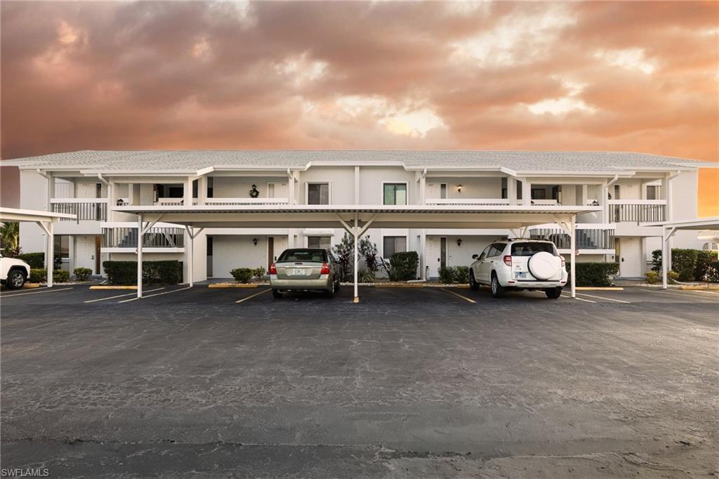Two-story residential building with light-colored exterior and multiple balconies, featuring covered parking structures