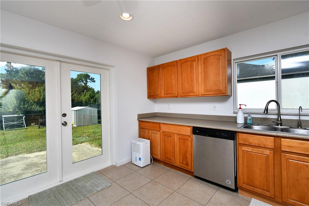 Wood-finish cabinetry, stainless steel dishwasher, dual basin sink with a gooseneck faucet, light-toned countertop, and tile flooring