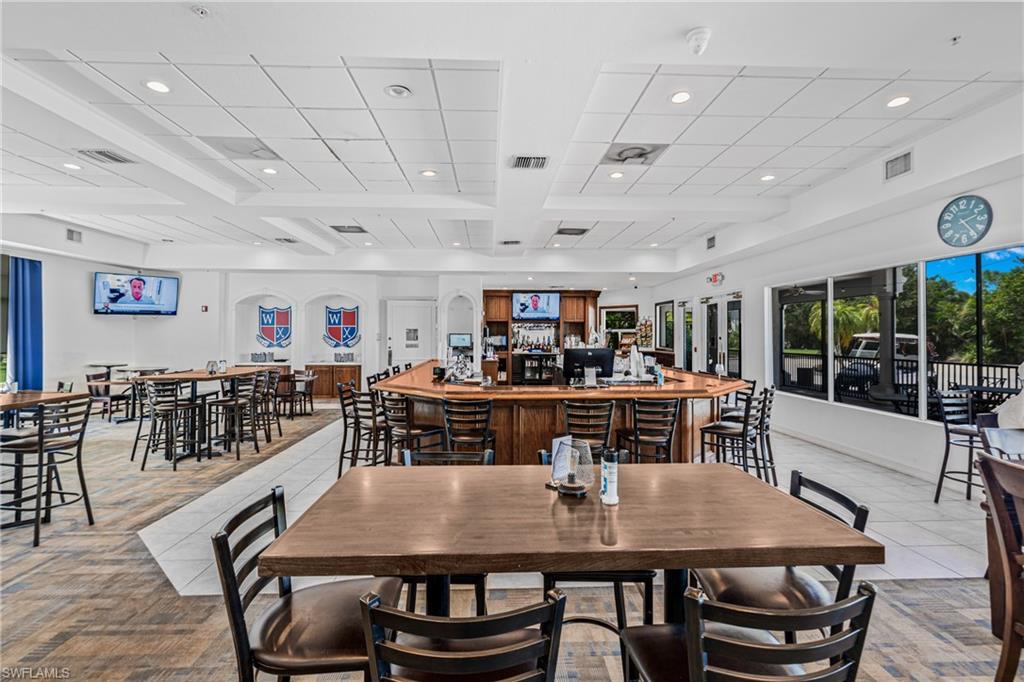 Expansive communal space featuring varied height tables with chairs, a built-in bar, wood-finish flooring, and large exterior windows