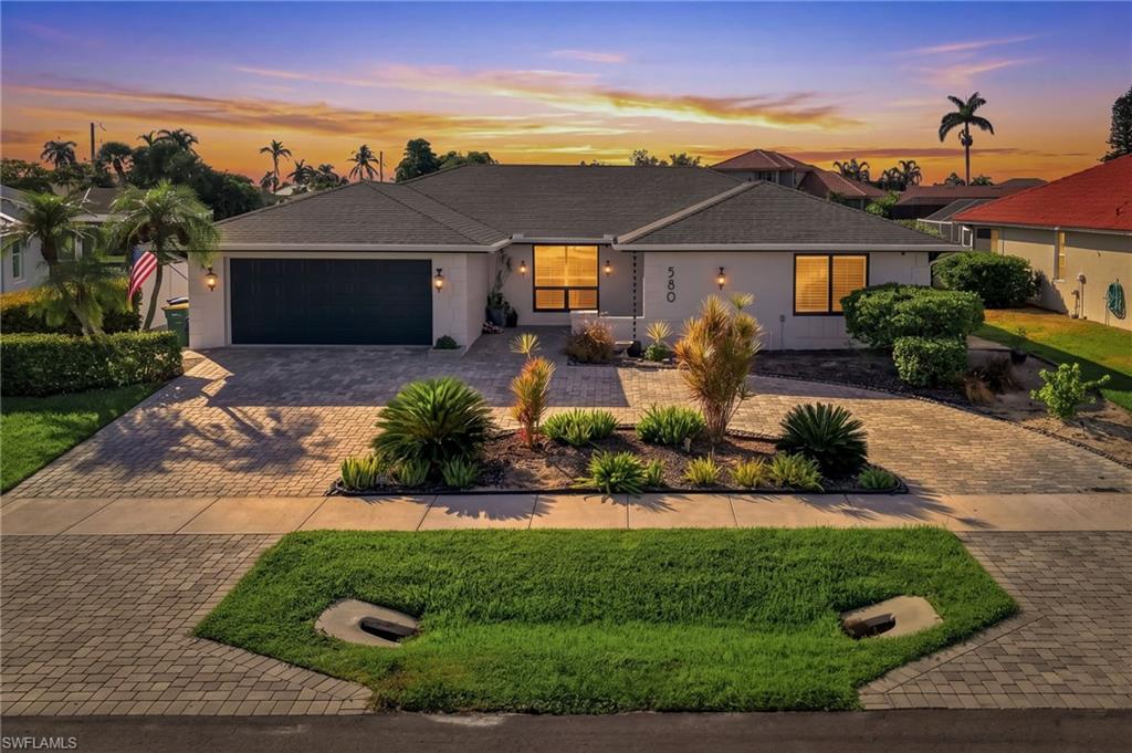 Paver driveway and walkway, mature landscaping, dark-toned garage door, exterior wall sconces, large windows with dark frames
