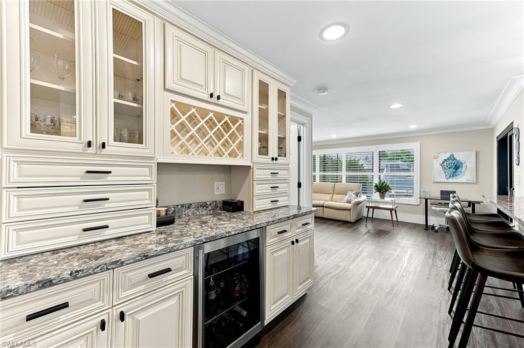 Cream cabinetry with glass fronts and black hardware, built-in wine rack, stone-finish countertops, undercounter beverage refrigerator, and wood-finish flooring