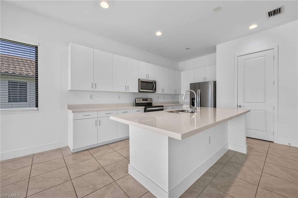 White cabinetry with brushed nickel hardware, stainless steel appliances, a large center island with an integrated sink, light-toned countertops, and neutral tile flooring