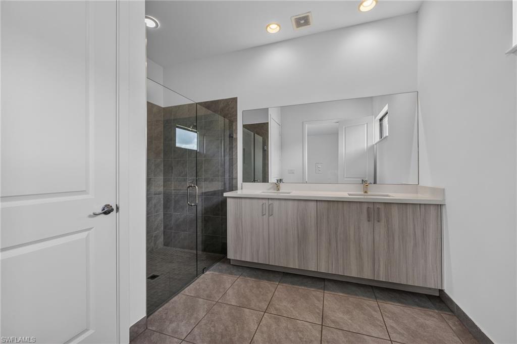 Dual vanity with light-tone wood-finish cabinetry, a full-width mirror, and a glass-enclosed shower with dark-toned tile