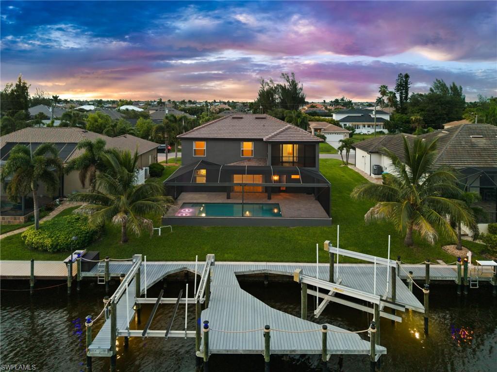 Striking rear waterfront view of the two-story home with screened pool enclosure, manicured backyard, custom dock, and two boat lifts. The upgraded waterfront setup includes remote-controlled 10,000-lb. and 16,000-lb. lifts for convenient boat storage at home.