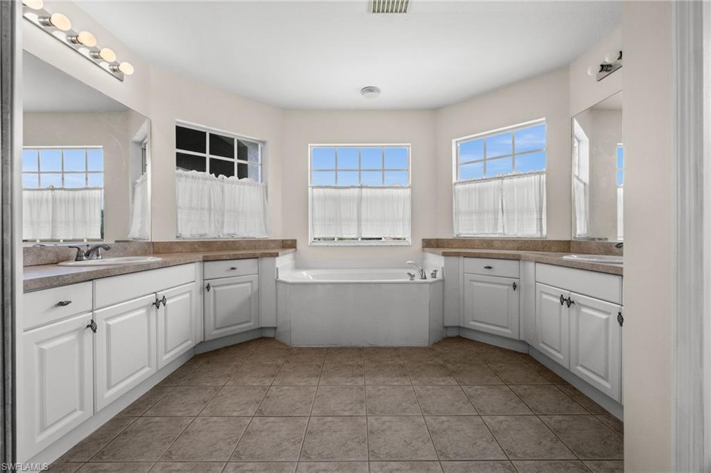 Primary bathroom offers dual vanities, abundant cabinetry, tile flooring, and a large soaking tub positioned beneath multiple windows. The spacious layout provides separate grooming areas and plentiful natural light.