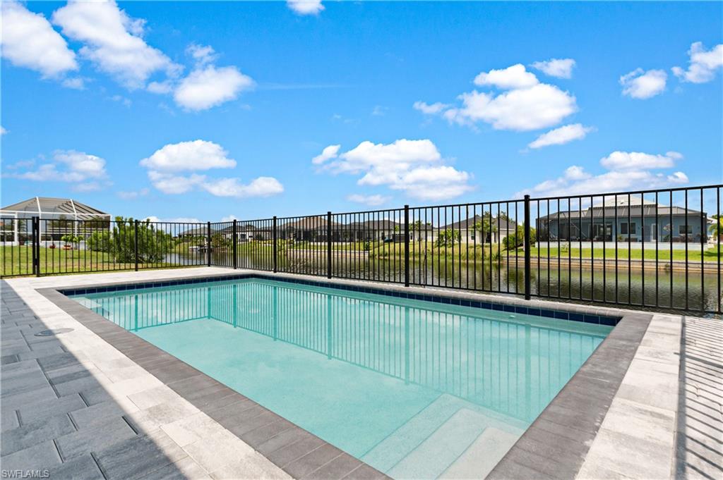 Outdoor swimming pool with integrated steps, surrounded by paver decking and a black metal fence, overlooking a body of water