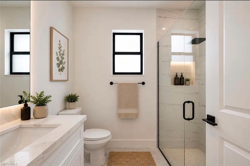 White vanity with an integrated sink and countertop, framed mirror with integrated lighting, glass-enclosed shower with a built-in niche, black-framed window, and matte black fixtures