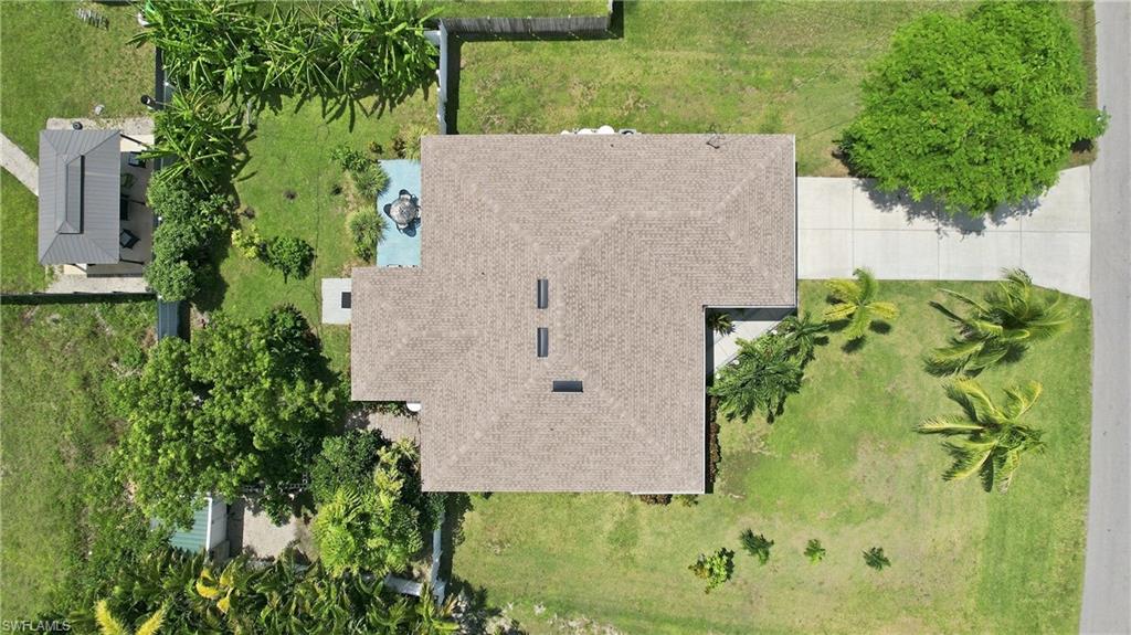 Pitched roof structure with multiple skylights, expansive green lawn, mature trees, concrete driveway, and an outdoor patio