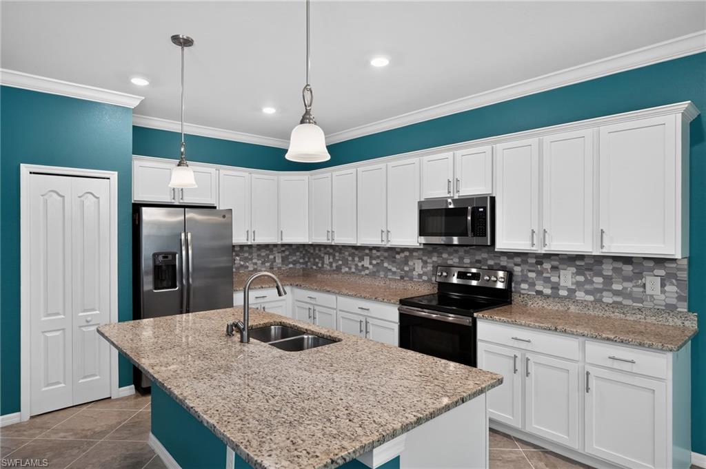 Contemporary kitchen featuring white cabinetry, granite-style countertops with waterfall island edge, stainless steel appliances including side-by-side refrigerator and dual wall ovens, glass tile backsplash, pendant lighting, and teal accent walls complementing the open layout