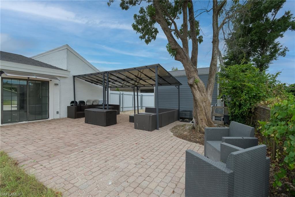 Expansive paver patio with a metal frame pergola, mature trees, and a sliding glass door entry to the building