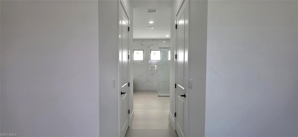 Recessed lighting illuminates a hallway leading to a bathroom featuring a frameless glass shower enclosure, three square windows, and a tiled accent wall