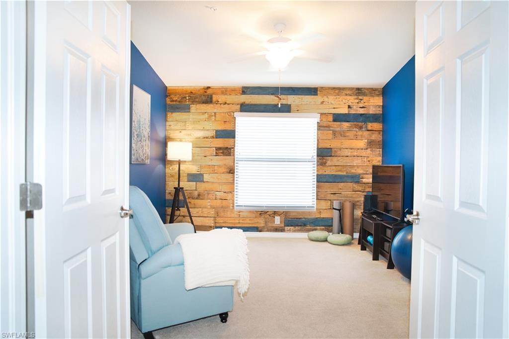 Textured wood accent wall, broad window with blinds, ceiling fan with illumination, carpeted flooring, and paneled interior doors