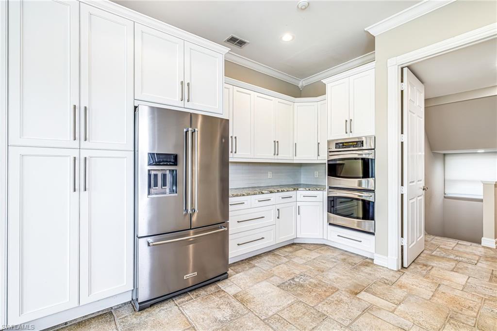White shaker cabinetry, stainless steel appliances, stacked wall ovens, natural stone-finish tile flooring, and granite countertops