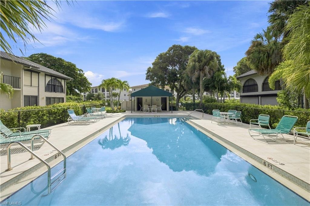 Expansive outdoor pool with a surrounding paved deck, lush perimeter landscaping, and a covered cabana area